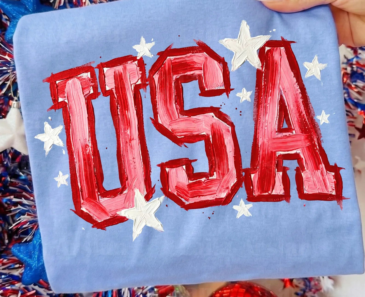 Blue pillow with red 'USA' text and white stars, held by a hand with a blurred background of festive decorations.