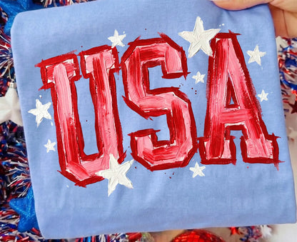 Blue pillow with red 'USA' text and white stars, held by a hand with a blurred background of festive decorations.